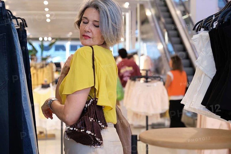 Mature woman with grey hair wears a bright yellow top and holds a stylish handbag while browsing in a modern clothing store. She appears thoughtful and relaxed