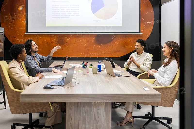 A diverse team collaborates in a modern office setting. They're engaged in a lively discussion around a large table with laptops, a whiteboard, and a projected presentation