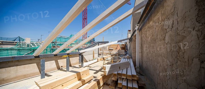 A construction site features exposed wooden beams and various building materials under a clear blue sky. Structural elements and scaffolding indicate progress