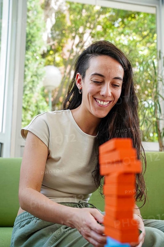 A young woman smiles while carefully playing a stacking block game indoors. The colorful tower and natural light create a warm, playful, and engaging atmosphere