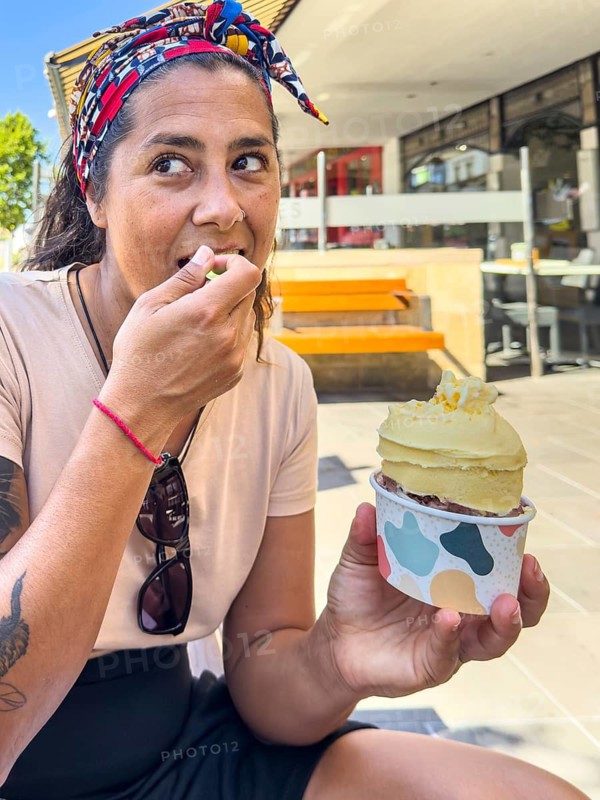 A woman seated outdoors savors a cup of ice cream. She dressed casually with a colorful headband, enjoying a sunny day in Victoria, Australia. The scene evokes a laid back, cheerful atmosphere