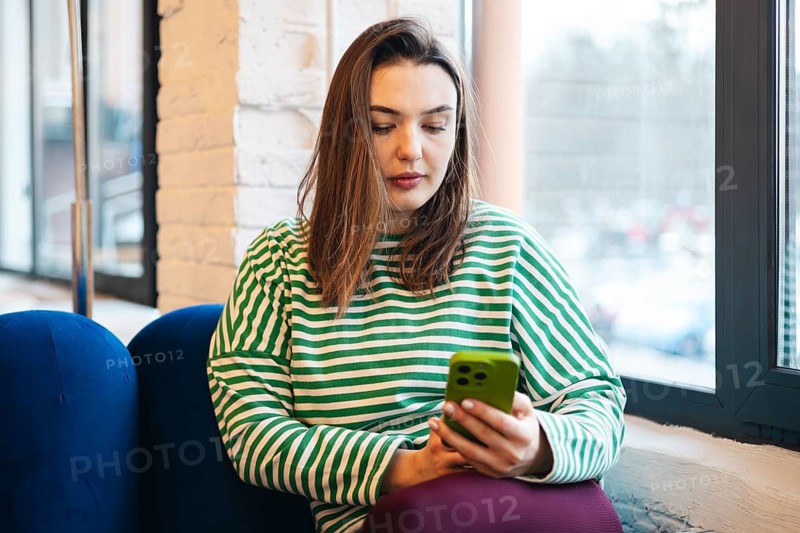 Woman in a striped shirt engrossed in her smartphone while sitting in a cozy cafe. The image captures a casual, relaxed moment during a coffee break