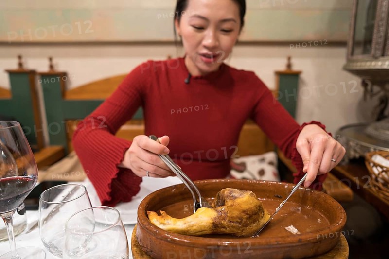 An Asian female tourist relishes a traditional roast suckling pig dish in a Segovian restaurant. She uses cutlery, showcasing her cultural exploration in Spain