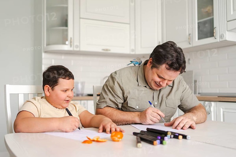 A father and son enjoy drawing together at the kitchen table, surrounded by art supplies and fresh fruit, while a small pet bird perches on the father shoulder