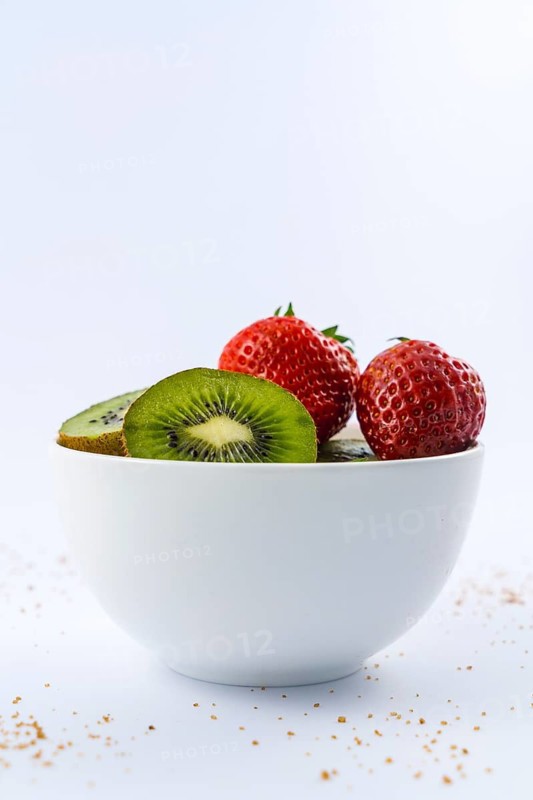 Low view of cut kiwis and strawberries in a white bowl with a white background with brown sugar
