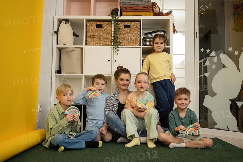 A teacher with five smiling children holding colorful crafts in a classroom. Shelves with educational materials create a warm, inviting learning environment