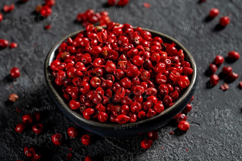 Top view of a dark bowl filled with bright red peppercorns scattered on a textured dark background, showcasing the vivid color and glossy texture of the spice