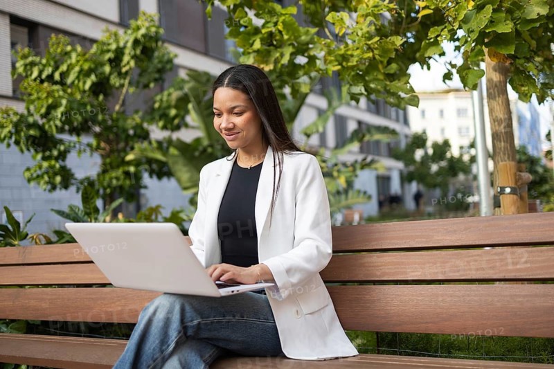 A Hispanic businesswoman in a white blazer works on her laptop while sitting on a bench in a green urban environment. She appears focused and professional