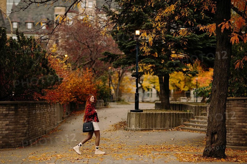 A woman strolls down a tree-lined path in Montreal, surrounded by vibrant autumn leaves. The scene captures the essence of fall with colorful foliage and a serene urban setting