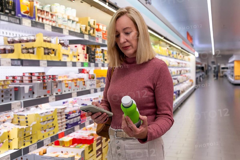 A woman in a supermarket aisle checks her smartphone while holding a green bottle, comparing product prices, showing modern shopping habits and decision making