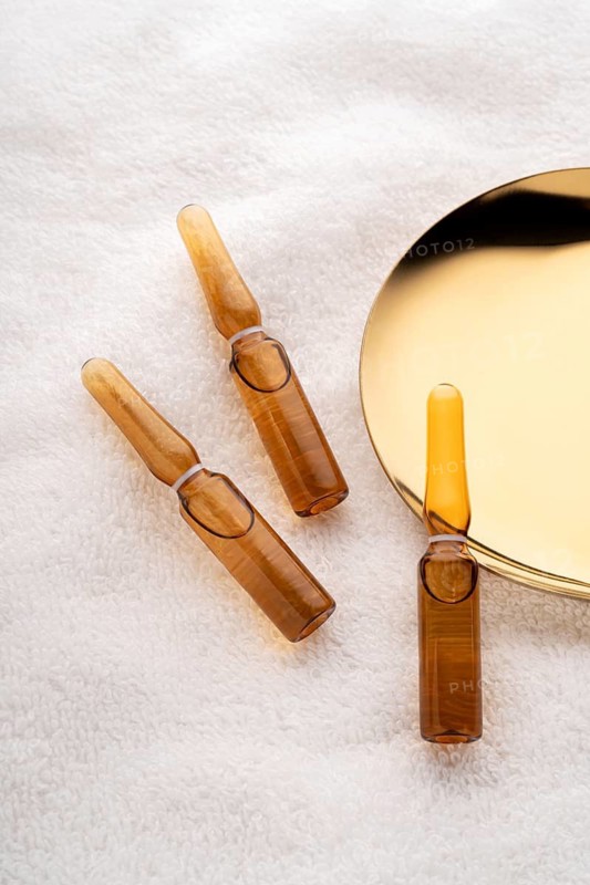 Three brown glass cosmetic ampoules filled with anti-aging serum elegantly displayed beside a reflective golden mirror on a white fluffy towel, emphasizing luxurious skincare routines