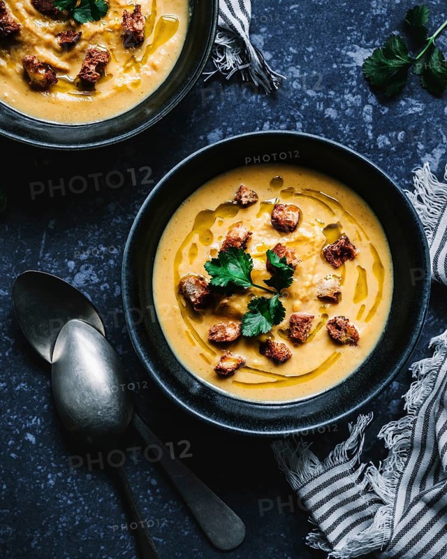 A rich, creamy soup served in a black bowl, garnished with crispy croutons and fresh parsley leaves, paired with a rustic cloth and metal spoons on a dark textured surface