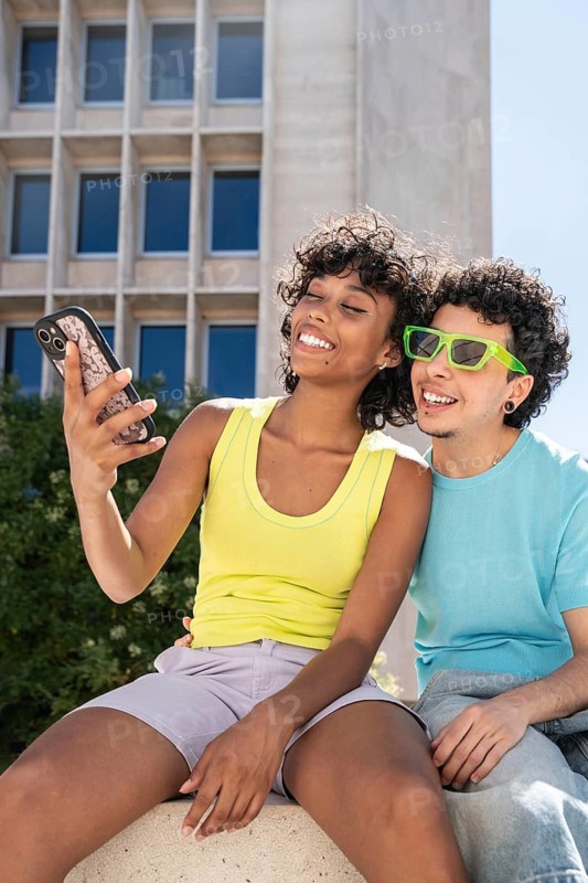 Smiling multiethnic LGBTQ+ couple in bright clothing taking a selfie outdoors with a modern building in the background. The pair radiate joy and love while enjoying their time together
