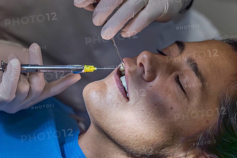 A dentist in gloves performs a teeth cleaning on a patient, administering anesthesia with a syringe. The patient is in a reclining position with eyes closed