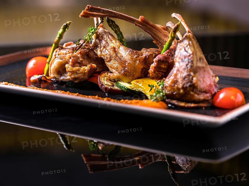 Gourmet lamb chops served with grilled vegetables like asparagus and tomatoes, elegantly plated on a black dish. Perfect for fine dining or culinary presentations