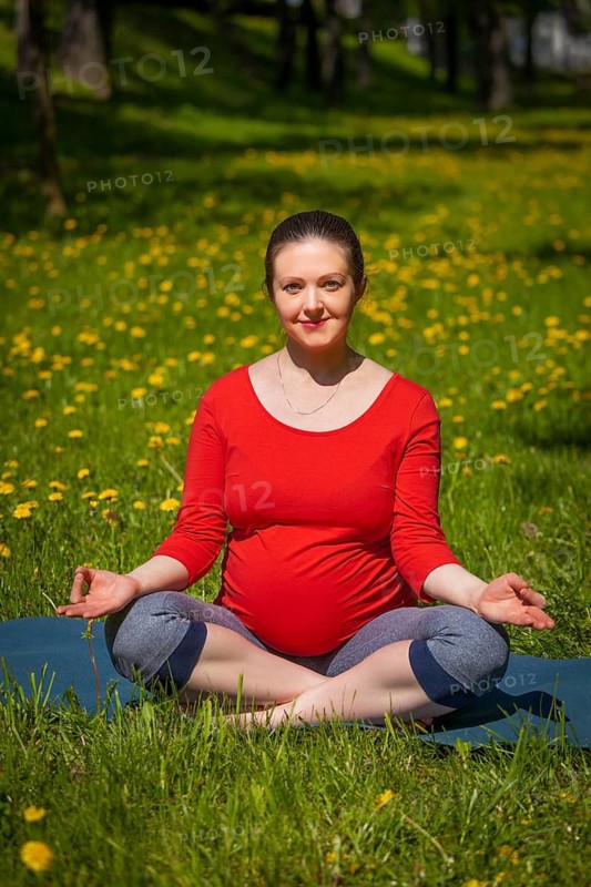 Pregnancy yoga exercise, pregnant woman doing asana Sukhasana easy yoga pose with chin mudra outdoors on grass lawn with dandelions in summer