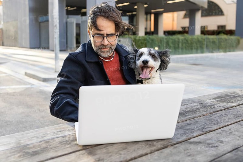 Man sitting outdoors with his dog, both focused on a laptop. This urban scene captures a casual yet engaging moment of pet companionship and mobile work, enhanced by the city environment