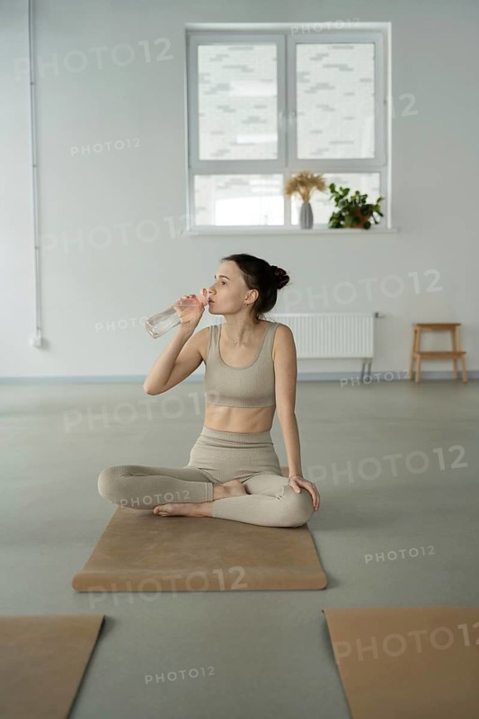 A woman in yoga attire sits cross legged on a mat, sipping water. The minimalist studio features soft lighting and indoor plants, creating a serene atmosphere
