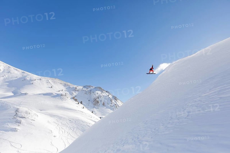 A snowboarder in mid air executing a jump against a backdrop of expansive snowy mountains. Clear blue sky enhances the exhilaration and freedom of the scene