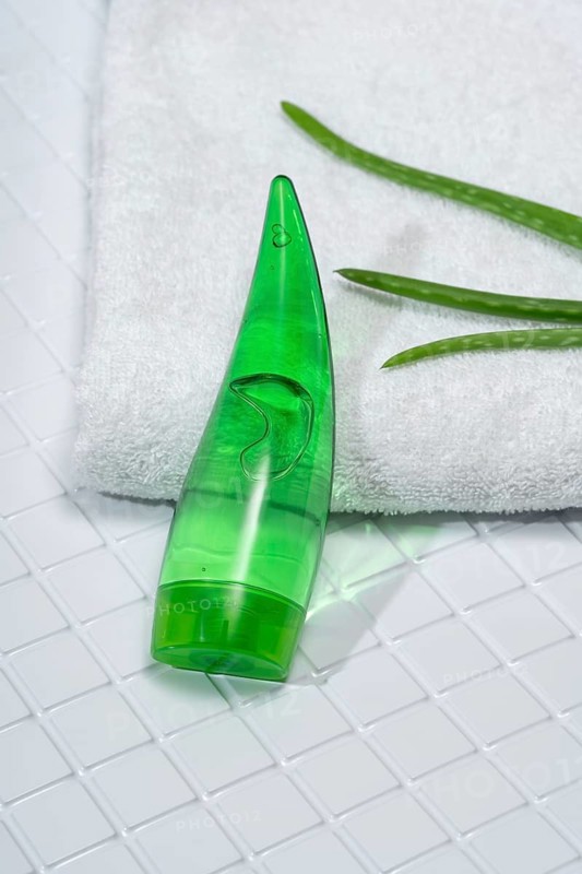 A transparent bottle of aloe vera gel rests on glossy white bathroom tiles alongside a fresh towel and aloe vera leaves