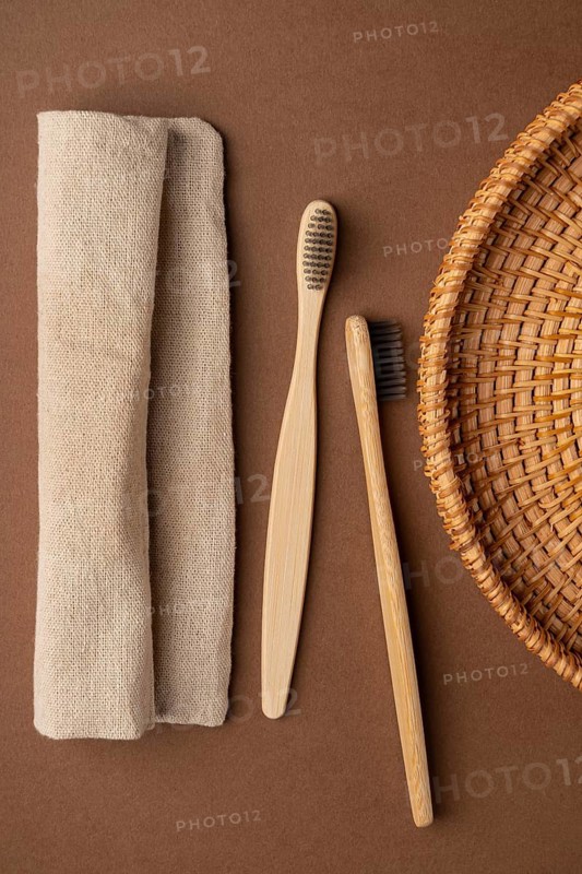 Top view of two bamboo toothbrushes alongside a beige linen towel and a wicker basket, set against a brown fabric background, showcasing eco-friendly toiletries