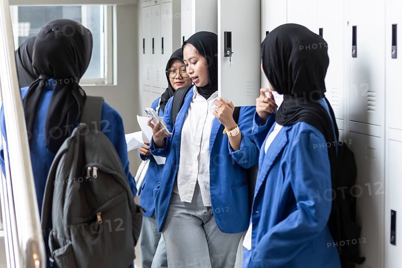 A group of Asian Muslim students in blue uniforms stand near lockers as they return to school. They are engaged with each other, using their phones and preparing for class