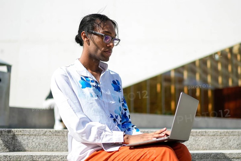 A thoughtful man wearing a floral shirt works on a laptop while seated outdoors. His attire and demeanor represent confidence, pride, and inclusivity in the LGBTQIA+ community