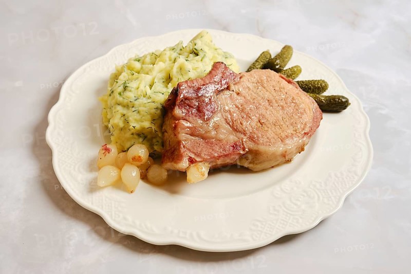 Roasted pork rib chop is placed on a plate next to creamy mashed potatoes and pickles. The dish is prepared for an evening meal. Onions are also present beside the meat and potatoes
