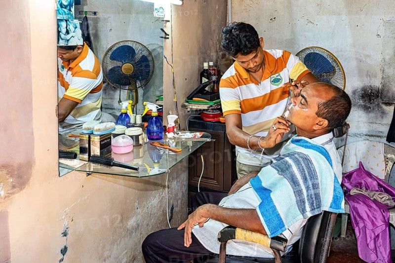 A hairdresser shaves a seated client in a simple room with a mirror and a fan, A man at a hairdresser in Sri Lanka