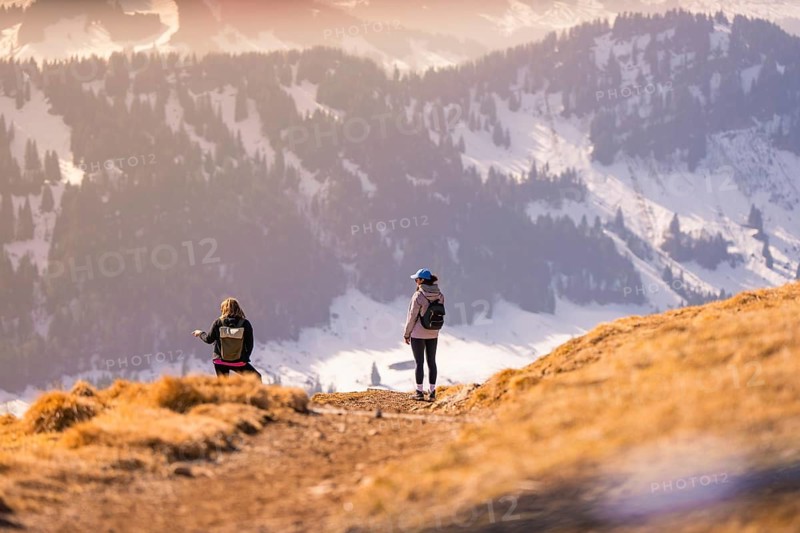 Two people hiking a sunny mountainside with snow-covered valley in the background, Hochgrat Mountains, Allgäu Alps, Oberreute, Germany