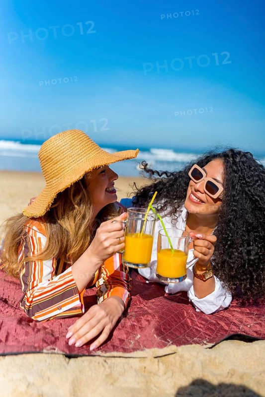 Two cheerful young women lying on a blanket on the sand, toasting with glasses of refreshing orange juice, celebrating their friendship and enjoying a relaxing summer day by the ocean