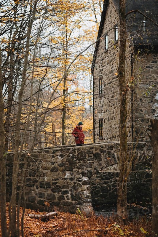 A woman walks across a stone bridge within a vibrant autumn forest in Quebec, Canada. The rustic stone structure complements the golden foliage of the surrounding trees