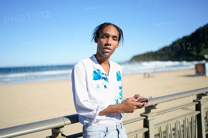 A confident man in a white shirt with blue accents stands by a beach railing, holding a smartphone. The sunny seaside view enhances the relaxed atmosphere