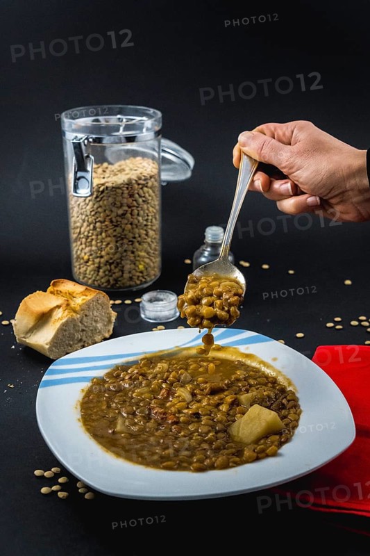 A hand of a young woman eating with a spoon inside a plate of Spanish cooked lentils, along with ingredients