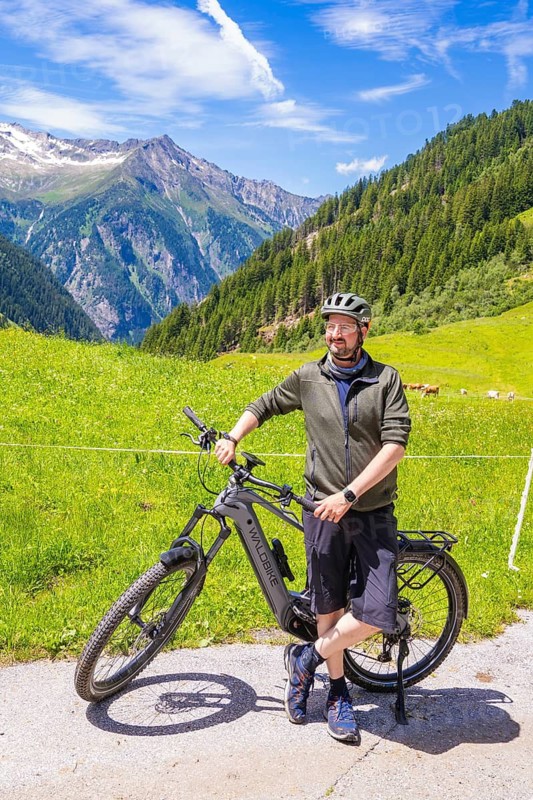 Man with helmet standing with his bicycle on a tarmac road, surrounded by green meadows and mountains under a blue sky, Klein Tibet, Zillertal, Austria