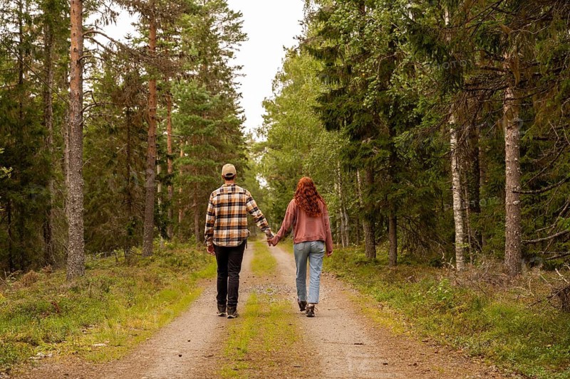 A couple walks down a serene forest path in Sweden, holding hands and enjoying nature. Tall trees surround them, creating a peaceful and romantic atmosphere