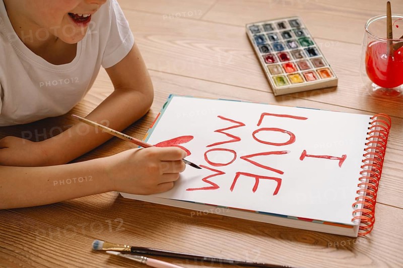 A young boy joyfully crafts a heartfelt Mother's Day card at home, using watercolor paint. The card expresses love and appreciation for his mom, showcasing creativity and affection