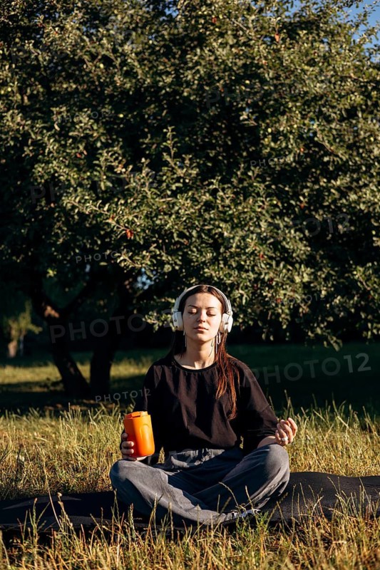 A teenage girl sits peacefully in a lush garden, eyes closed, wearing headphones, meditating or relaxing in the serene morning. The tranquility of the morning surrounds her