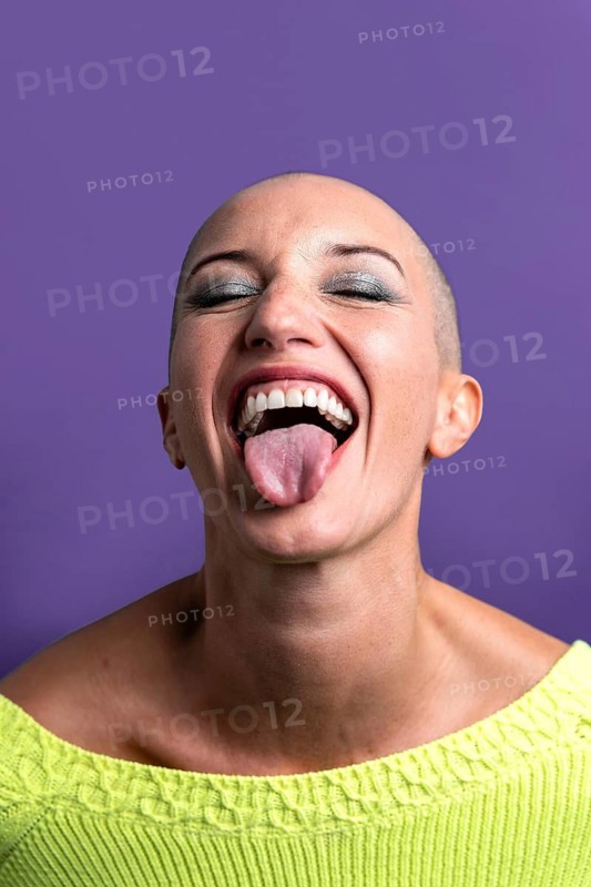 Middle-aged woman with a shaved head joyfully sticks out her tongue, conveying a sense of freedom and boldness. Her bright green sweater contrasts with the purple background
