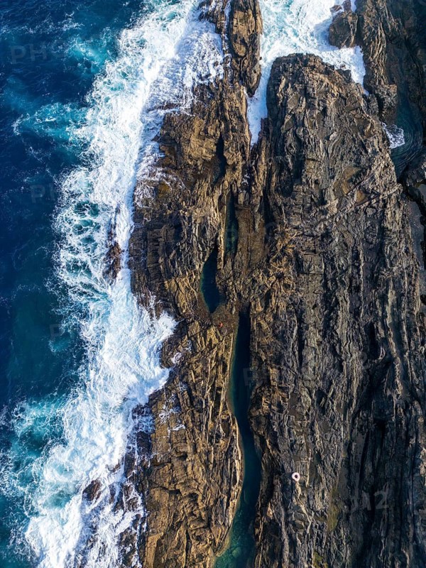 From above, captured by a drone, this image showcases the rugged beauty of Tindaya Beach in Fuerteventura, Canary Islands. The aerial shot highlights natural pools carved into the weathered rocks, surrounded by the white waves of the Atlantic