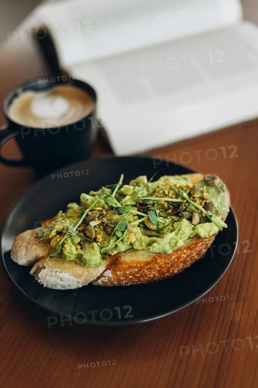 A plate of avocado toast topped with fresh greens and seeds, served alongside a steaming cup of coffee on a wooden table. A book is open in the background