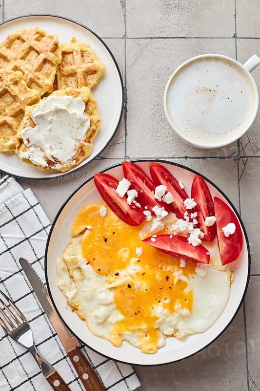 Top view of a delightful homemade breakfast featuring a fried egg topped with feta cheese and fresh tomatoes, alongside Belgian waffles covered in cream cheese. Completing the meal is a cup of coffee, perfect for a relaxing morning