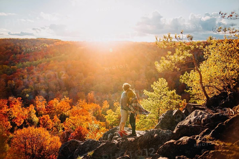 A couple embraces on a rocky overlook, surrounded by vibrant autumn foliage in Quebec, Canada. The sun sets, casting a warm glow over the brilliant fall colors