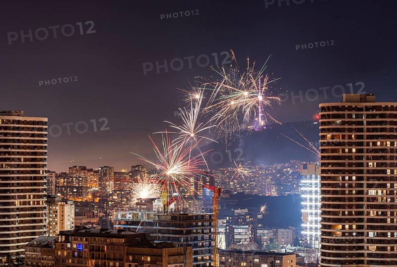 Night view of Tbilisi's downtown, showcasing a vibrant fireworks display above the cityscape. Tall buildings frame the scene, reflecting the lively atmosphere