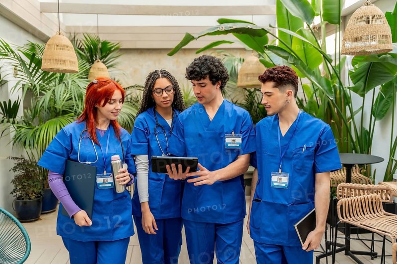 Healthcare students in blue scrubs and stethoscopes are collaborating and discussing information on a digital tablet, learning and working together in a calm, modern, and green study space