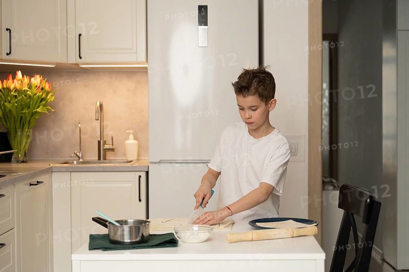A young boy focuses on preparing a meal in a modern kitchen. He uses a knife on a white counter with pans, bowls, and flowers in the background, creating a warm atmosphere