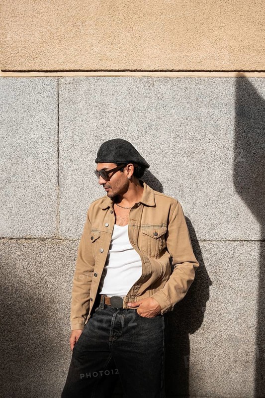 A man stands casually in front of a stone wall, wearing sunglasses, a flat cap, a beige jacket, and a white tank top. His relaxed pose and attire give off an urban vibe