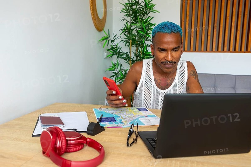 A blue-haired LGBTQIA+ man plans a holiday trip with a laptop and phone. Maps, headphones, and notes are scattered on a wooden table, creating a cozy atmosphere