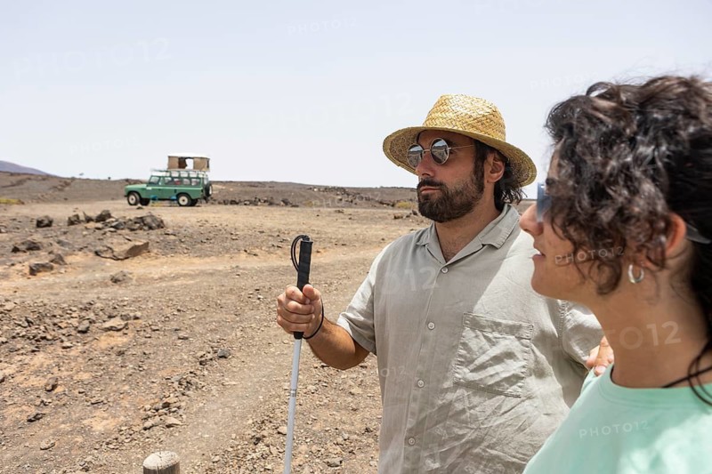 A blind man with a cane and a woman stand together in a desert landscape under the bright sun, enjoying the experience of exploration with a vintage off-road vehicle in the background