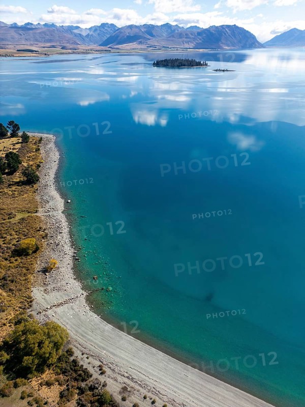 Aerial view of Lake Tekapo in Canterbury, New Zealand, showcasing a breathtaking turquoise lake with a small forested island, surrounded by a mountainous landscape